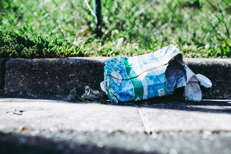 A crumpled, discarded plastic bag made of thin, glossy material in shades of blue and white, lying on a concrete pavement near the edge of a curb. The bag appears to be partially torn and is tangled with a black plastic tie or string. The background features a strip of green grass and plants, indicating an outdoor urban or suburban environment. The scene is well-lit with natural sunlight, casting shadows on the pavement and emphasizing the texture of the bag and surrounding concrete. The image depicts common roadside rubbish, highlighting the importance of proper waste disposal and the role of independent rubbish removal services such as Rubbish Clearance Brentford in managing waste that might otherwise be improperly discarded in public areas.