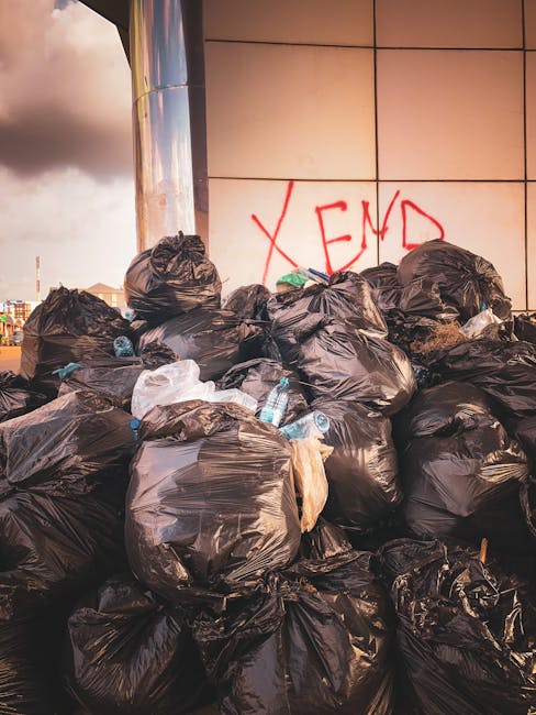 A large collection of tightly tied black plastic rubbish bags, stacked in front of a modern tiled wall with a graffiti message 'XEND' spray-painted in red. The bags, containing mixed waste including visible plastic bottles, occupy most of the foreground and are piled on a flat surface outside a building, indicating an accumulation of waste typical of a rubbish clearance scenario. The environment appears to be an urban exterior setting, with a cloudy sky and distant cityscape visible in the background. The scene emphasizes the need for waste removal services, such as those provided by Rubbish Clearance Brentford, as part of alternative waste handling or on-site clearance solutions to manage excess rubbish effectively in residential or commercial areas.