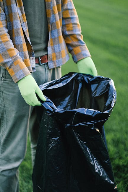 A person standing outdoors on a grassy area, wearing a plaid shirt over a dark T-shirt, with light green gloves on both hands, holding open a large black rubbish bag lined with a silver inner layer. The individual appears to be preparing the bag for waste collection. The background features a lush, green landscape with a blurred view of grass and possibly trees. This scene reflects an activity related to rubbish removal or waste clearance, potentially undertaken by a waste management service such as Rubbish Clearance Brentford, which specializes in private waste disposal or on-site rubbish collection. The focus on the waste bag and the gloves emphasizes a professional and environmentally conscious approach to rubbish collection, aligning with the context of independent rubbish clearance services.