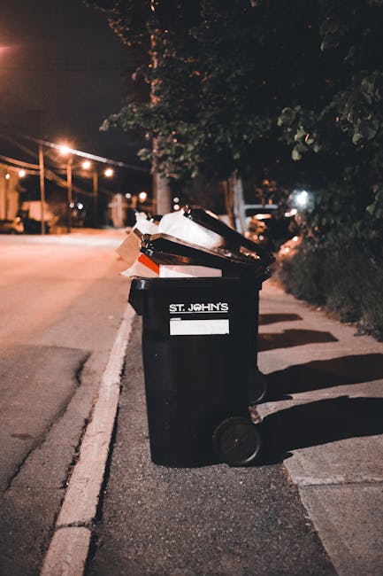 A black wheelie bin with a white label reading 'St. John's' positioned on a sidewalk next to a curb in an urban area at night. The bin is filled with various types of rubbish, including cardboard boxes, black plastic lids, and light-colored packaging material, which are visible as they protrude from the open top. The surrounding environment includes a street illuminated by streetlights, with a background of trees and distant parked cars. The pavement appears clean, and the scene suggests a private rubbish collection or on-site waste clearance service provided by Rubbish Clearance Brentford. Shadows cast by the bin and nearby trees are visible on the ground, emphasizing the nighttime setting, while the dim ambient lighting highlights the focus on the bin and its contents, reflecting typical waste management scenes in urban residential or commercial areas.