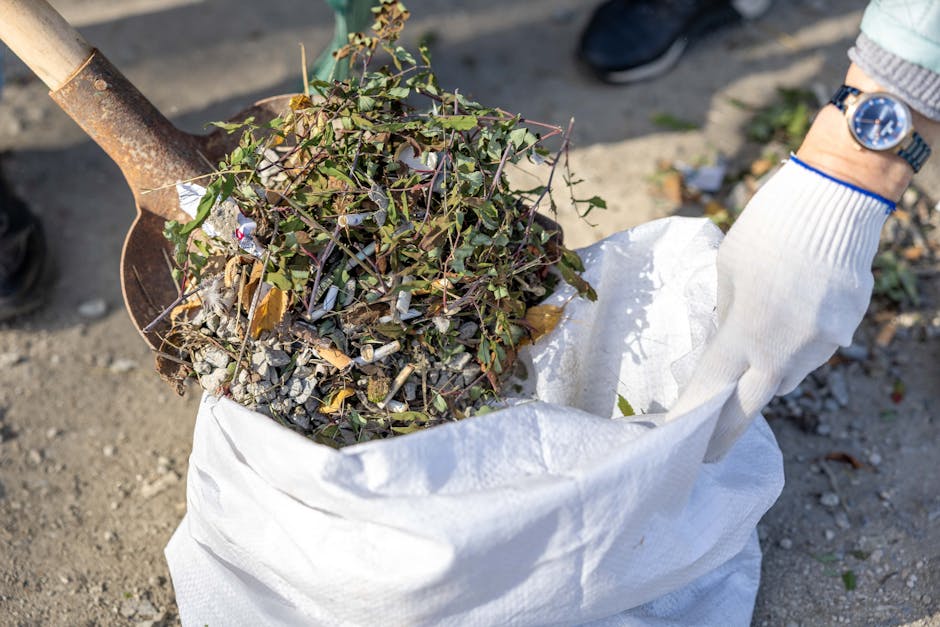 A person wearing a white work glove and a silver wristwatch is using a rusted metal shovel to transfer a mixture of garden waste, including small branches, leaves, and twigs, into a large white woven polypropylene sack. The garden debris appears fresh with green and brown foliage, some twigs with bark, and small bits of gravel or soil mixed in. The scene is set outdoors on a gravel or dirt surface, with sunlight casting natural shadows, indicating daytime. The person's arm is visible, wearing a long-sleeved light-colored garment, while the background shows a blurred outdoor environment. This visual relates to onsite or private waste clearance activities, typical for garden or green waste removal services provided by Rubbish Clearance Brentford, supporting their reputation for independent rubbish removal and disposal solutions near Brentford Station, with an emphasis on environmentally responsible rubbish handling. The image captures a moment of waste collection aligned with alternative rubbish removal options outside of local authority collections.